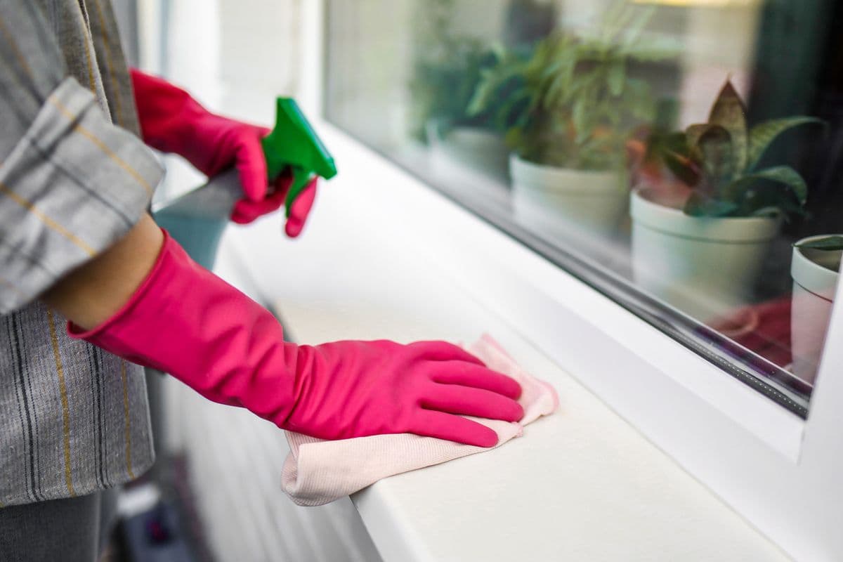 Gloved hand spraying and wiping a white windowsill